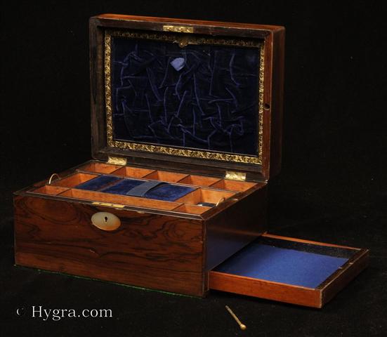 Antique Figured Rosewood box with lift-out compartmentalized tray and sprung drawer fitted for jewelry. There are mother of pearl escutcheons to the top and the front. There is a ruched velvet pad in the lid is framed with gold embossed leather behind. It is released by pressing behind the lock catch. The sprung drawer is released by removing a brass pin. The lift-out tray is of mahogany construction with rosewood facings. New pads of velvet have been made to make the tray suitable for jewelry. There is further space beneath. Circa 1840. - Enlarge Picture